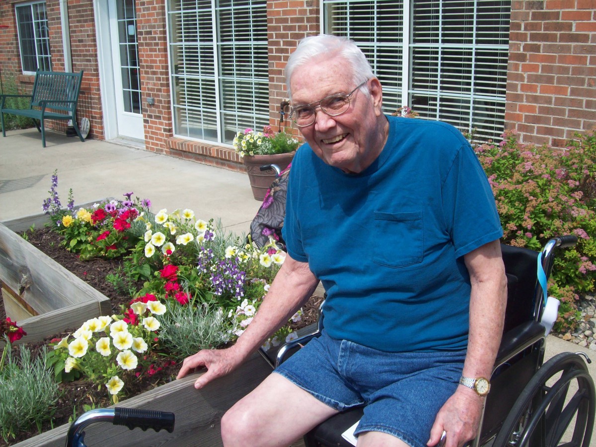 An elderly man sitting by a garden full of colorful flowers.
