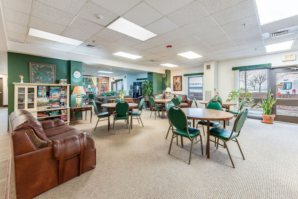 A spacious community room with tables and chairs, decorated in green tones, featuring indoor plants and shelves.