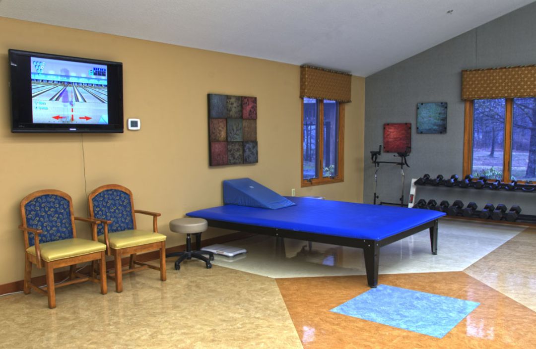 A well-equipped physical therapy room with a therapy bed, exercise equipment, and chairs.