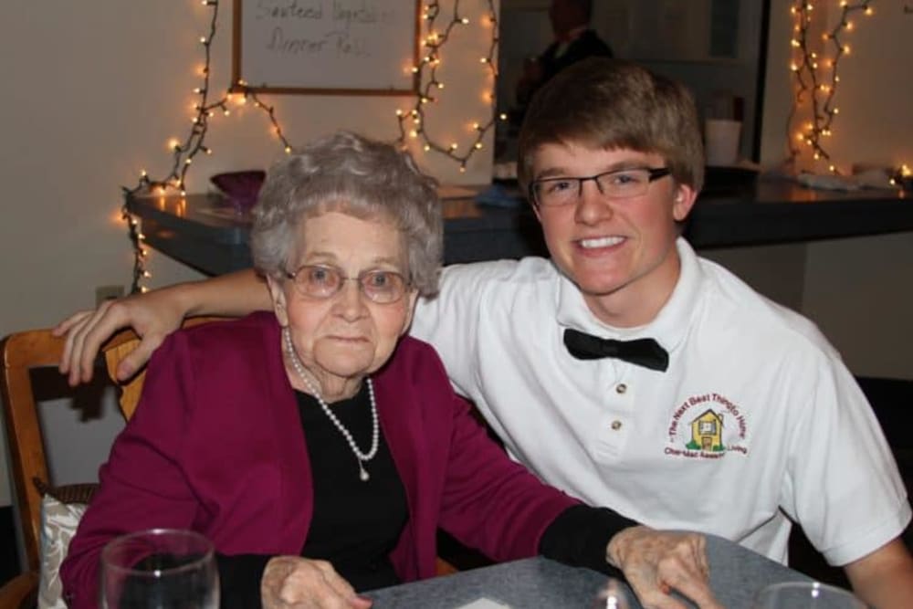 A young man with glasses poses with an elderly woman, both smiling warmly in a cozy indoor setting.