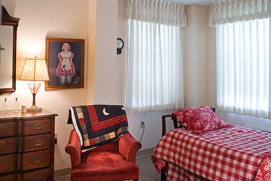 A cozy bedroom with a red armchair, a quilt on the bed, and a small lamp on a wooden table next to a painting.
