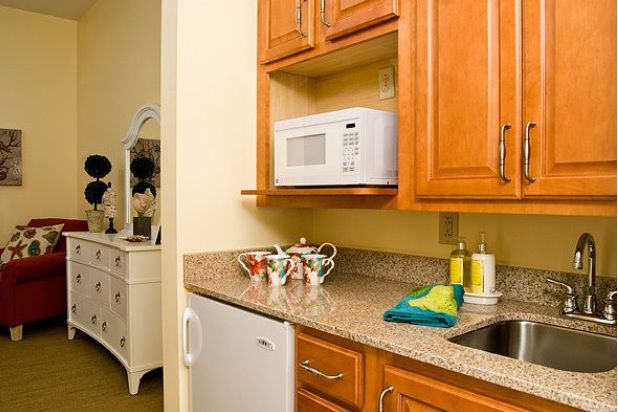 A cozy kitchen corner featuring a microwave within wooden cabinets, a small fridge, and teacups on the countertop.