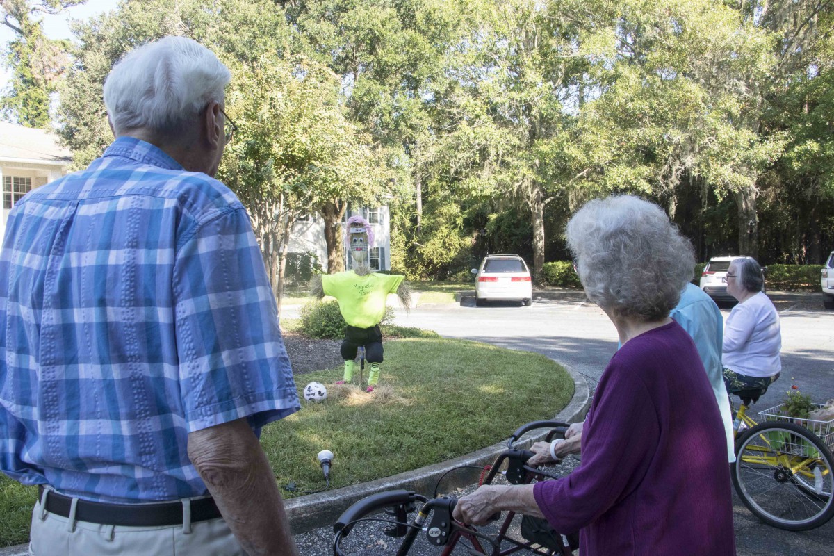 An elderly couple watches a person in reflective clothing working in a neighborhood garden.