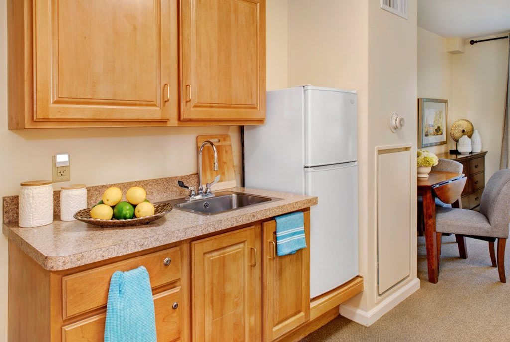A kitchen with wooden cabinets, a countertop with a sink, and a refrigerator. Towels are hung, and a fruit bowl is on the counter.