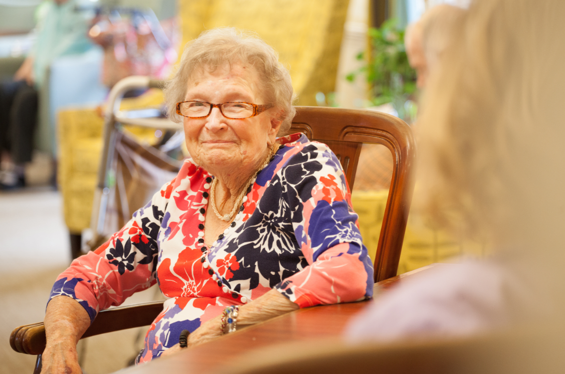 An elderly woman wearing glasses and a colorful dress sits comfortably indoors, smiling warmly.