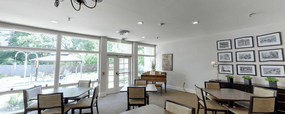 A bright room with a piano, tables, and chairs, illuminated by natural light from large windows.