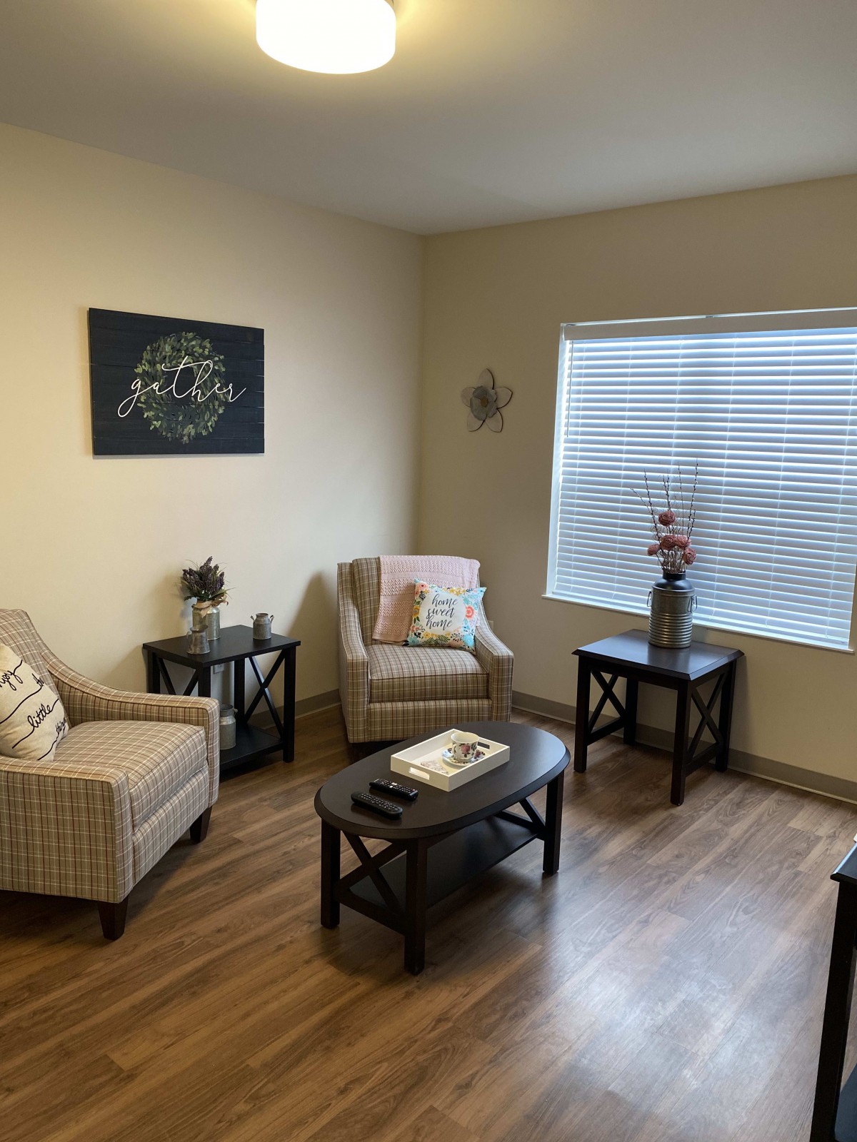 A cozy living room with armchairs, a coffee table, and side tables, featuring wooden floors and natural light from a large window.