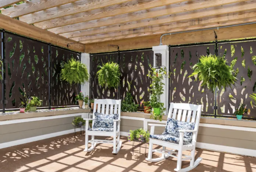 A cozy outdoor patio featuring two white rocking chairs surrounded by lush greenery.