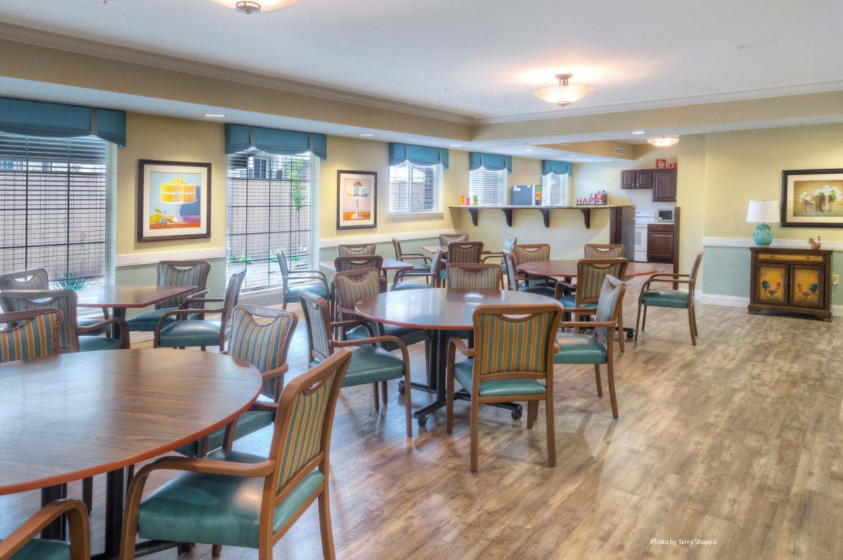 A bright and spacious dining area with wood flooring, featuring multiple tables and chairs, decorated with artwork on the walls and illuminated by ceiling lights.