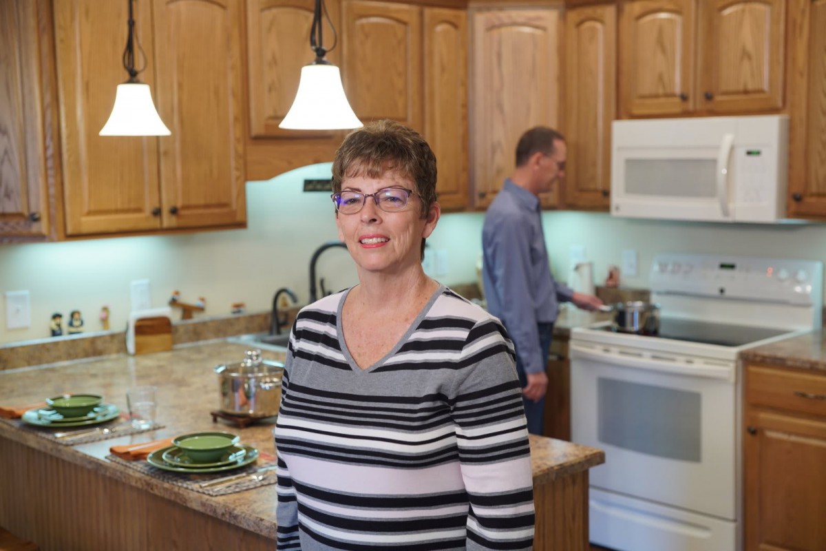 A woman smiling in the kitchen while a man is cooking in the background.