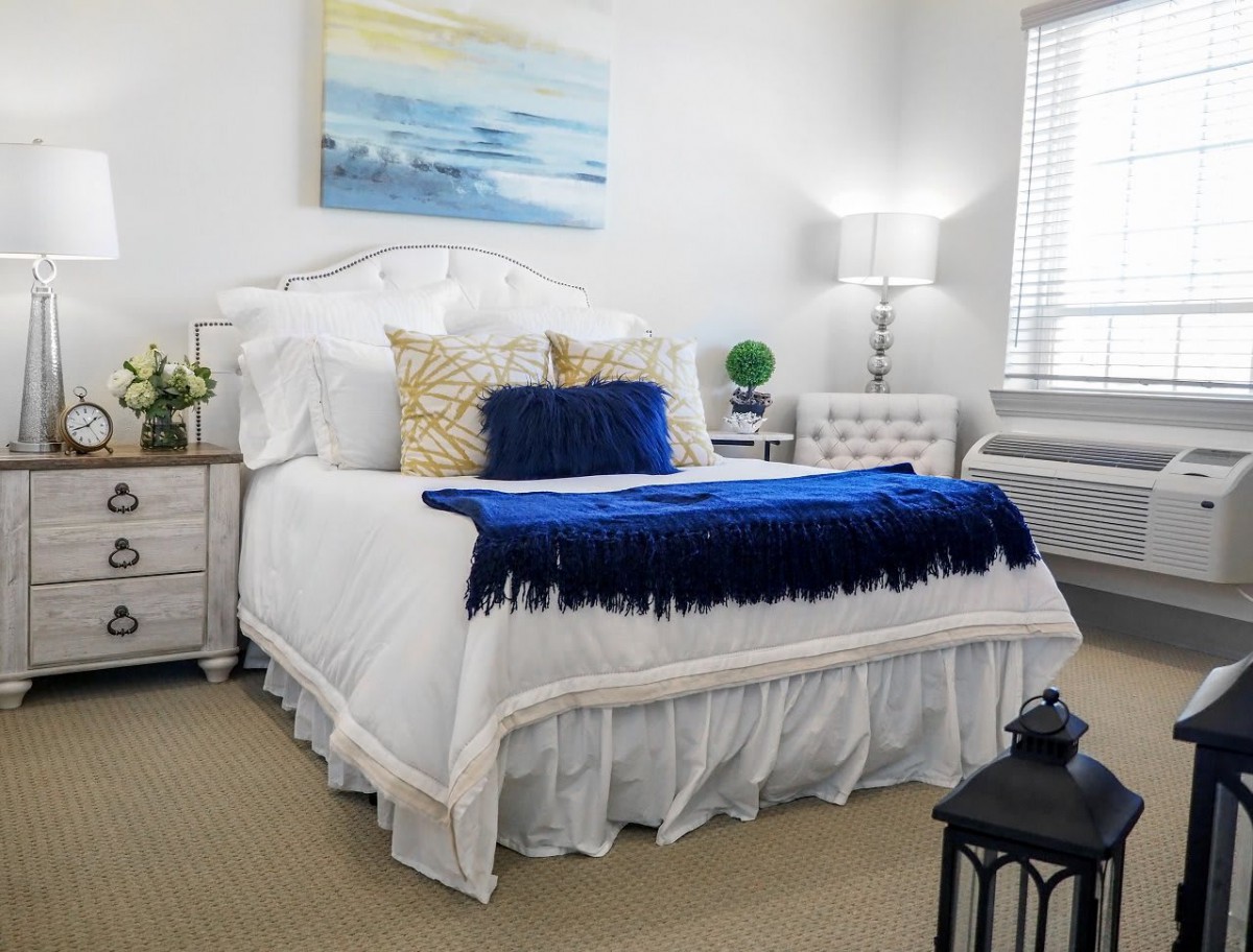 A cozy bedroom with a neatly made bed featuring blue and white bedding, accented by decorative pillows. A lamp and artwork add to the room's charm.