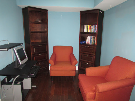 A cozy office space featuring two orange chairs, dark wooden bookshelves filled with books, and a computer setup.
