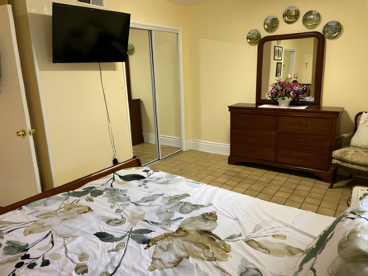 A cozy bedroom with a wall-mounted TV, mirrored closet, and wooden dresser adorned with decorative plates.