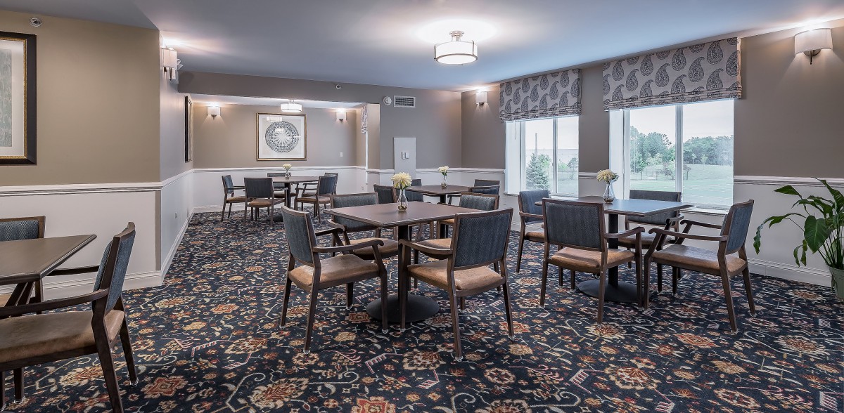 A well-lit dining room with several tables and chairs arranged on a patterned carpet.