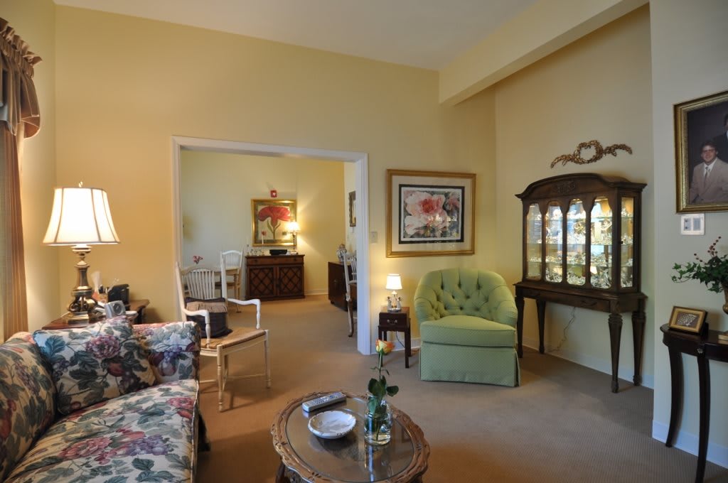 A cozy living room with traditional decor, featuring a floral sofa and green armchair, complemented by a wooden cabinet and artwork on the walls.