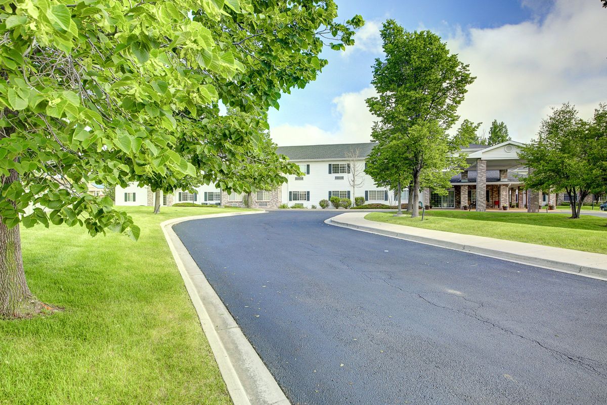 A quiet driveway leading to a white building surrounded by lush greenery on a sunny day.