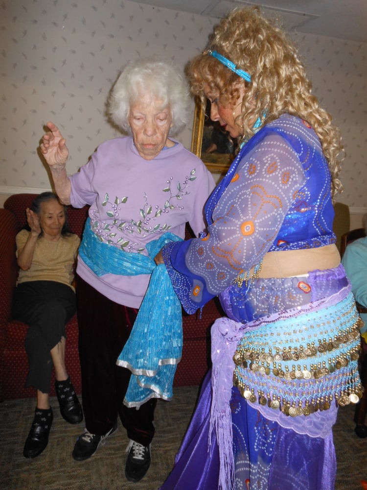 An elderly woman and a belly dancer in a colorful costume share a moment as they dance together indoors.