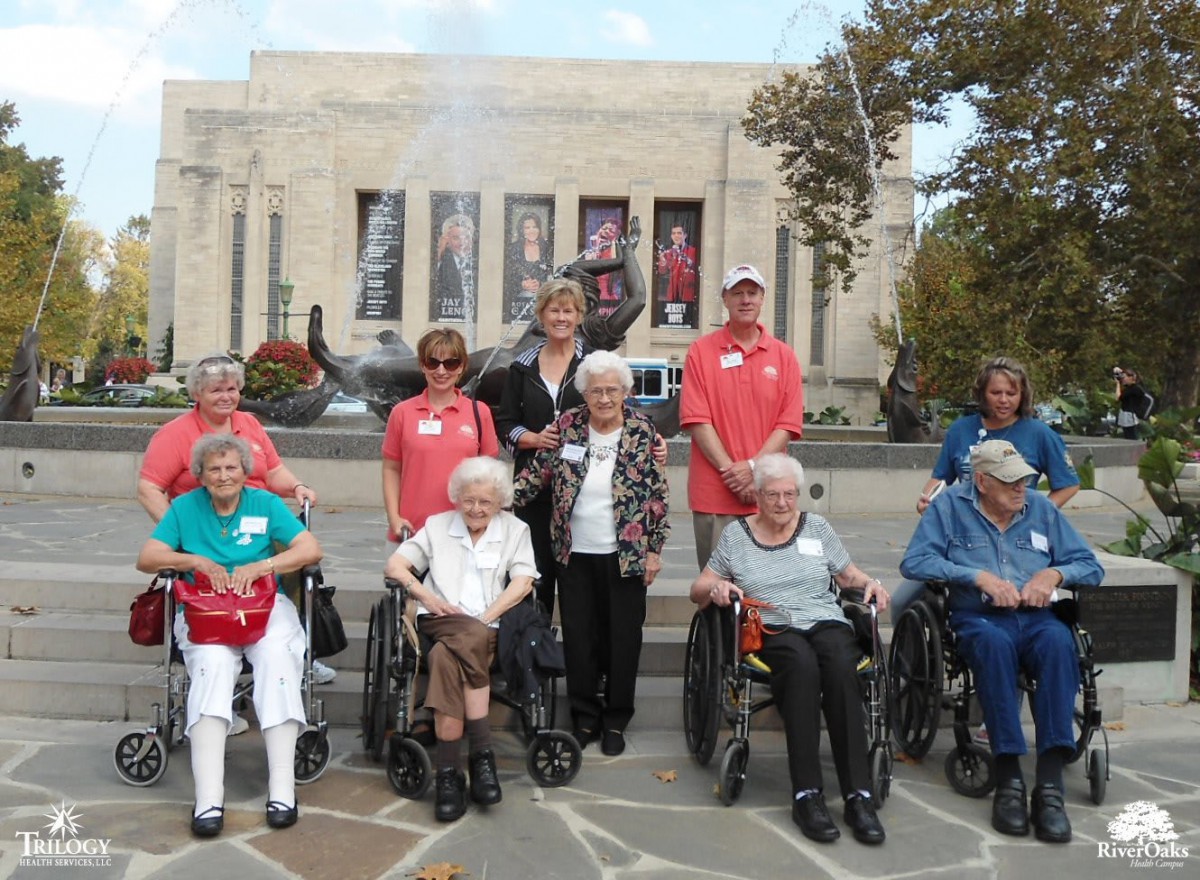 A group of seniors, some in wheelchairs, pose for a photo in front of a large building during the day.