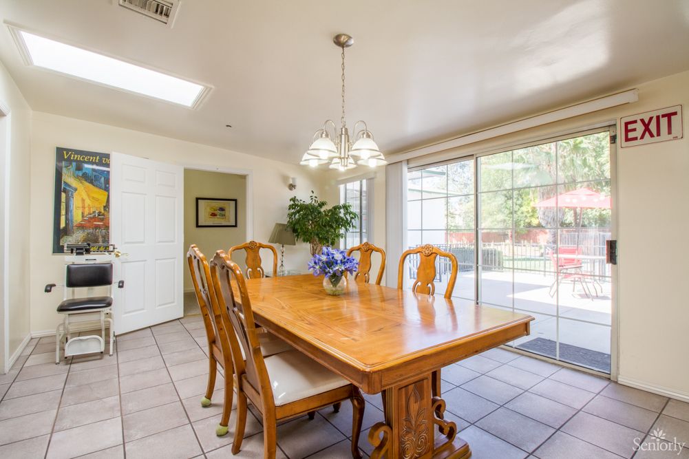 A bright dining room with a wooden table and chairs, illuminated by a chandelier, featuring an indoor plant and a sliding glass door.