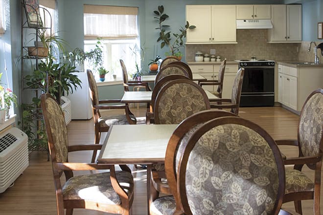 A cozy dining area with elegant chairs and tables, illuminated by daylight from the windows, featuring a kitchen in the background.