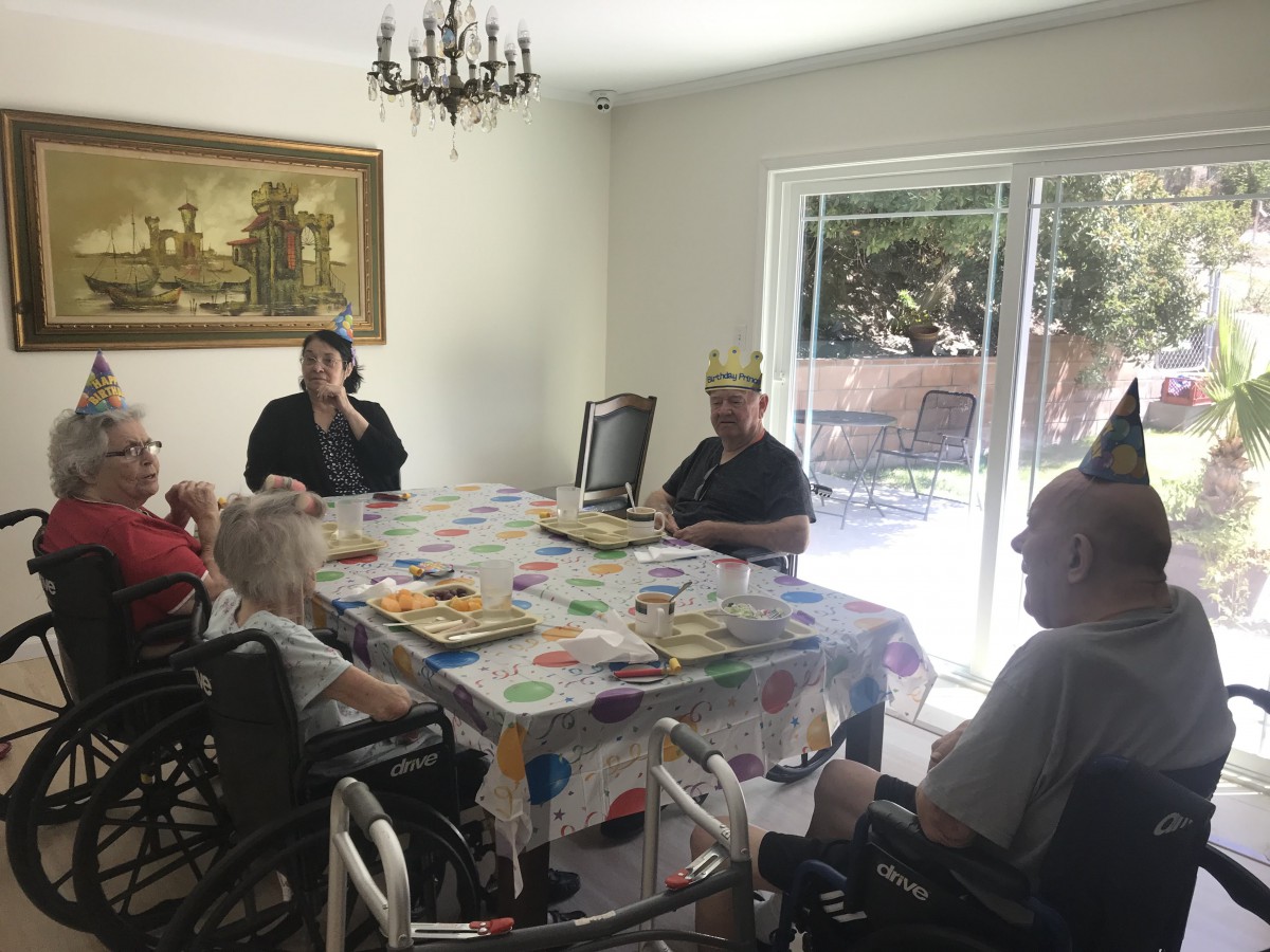 A group of elderly people gathered around a dining table, celebrating with a birthday party. One person is wearing a crown, suggesting they are the guest of honor.