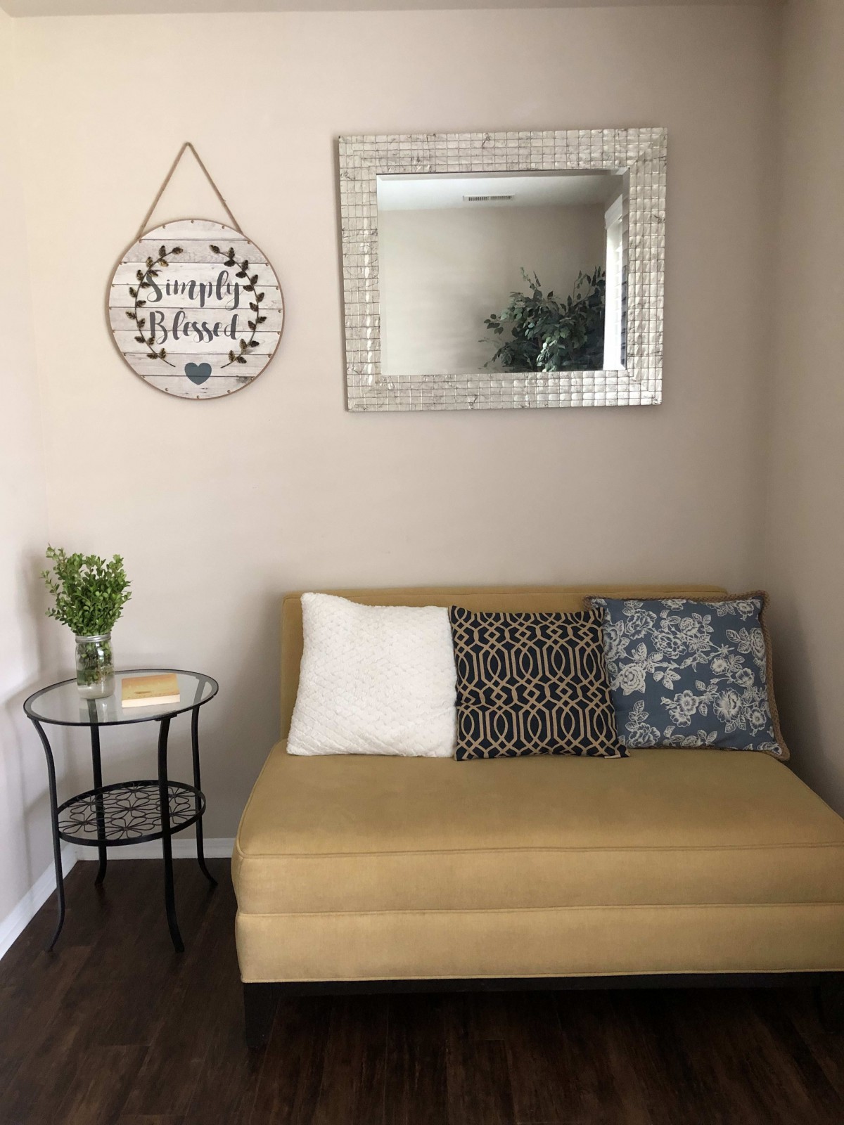 A cozy living room corner with a tan sofa, decorative pillows, a round side table with a vase of flowers, and a stylish wall mirror.