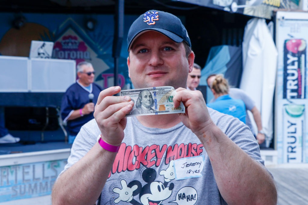A man wearing a baseball cap holds up a $100 bill while standing outdoors.