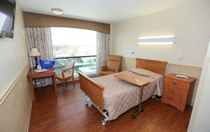 A clean and organized hospital room with a single bed, a chair, and a window providing natural light.