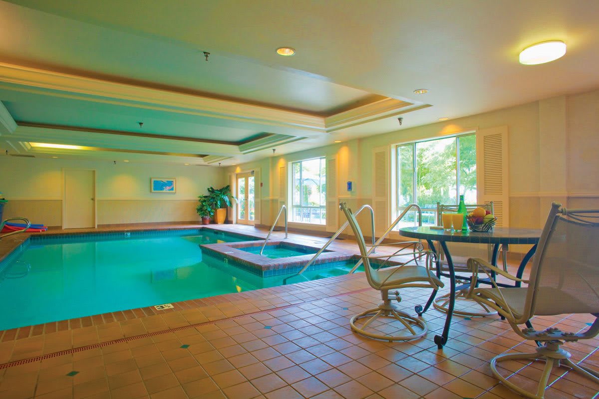 A bright indoor swimming pool area with chairs and a table by the window, offering a relaxing environment.
