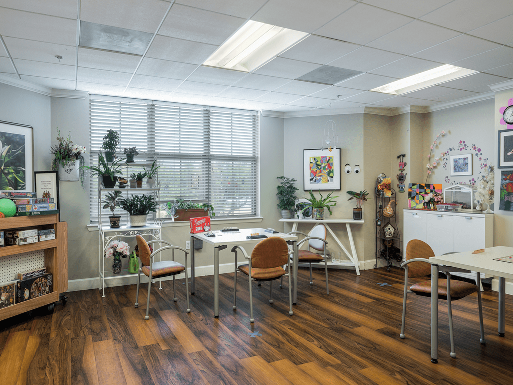 A modern office interior with large windows, decorated with plants and featuring desks and chairs on a wooden floor.