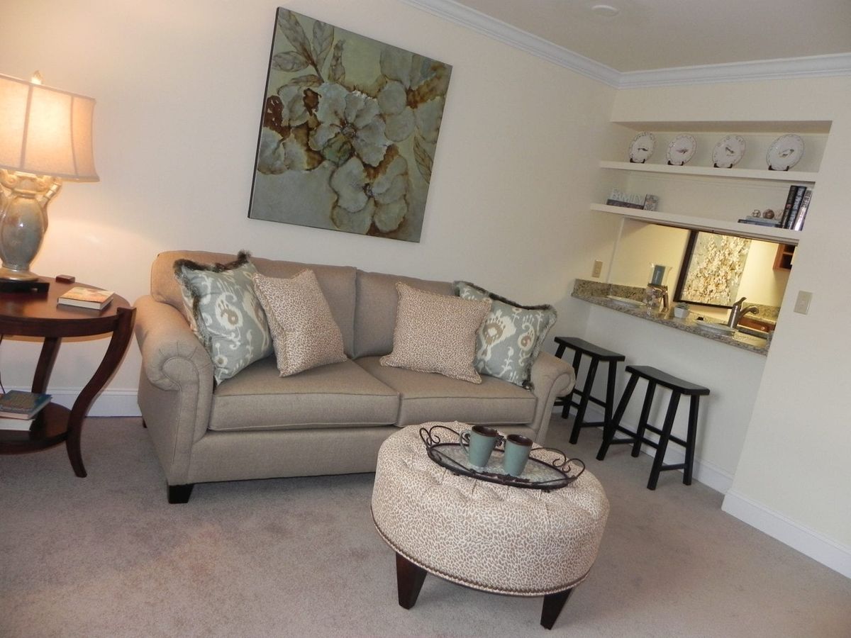 A cozy living room with a beige sofa and an ottoman, accented by a piece of artwork on the wall and bar stools in the corner.