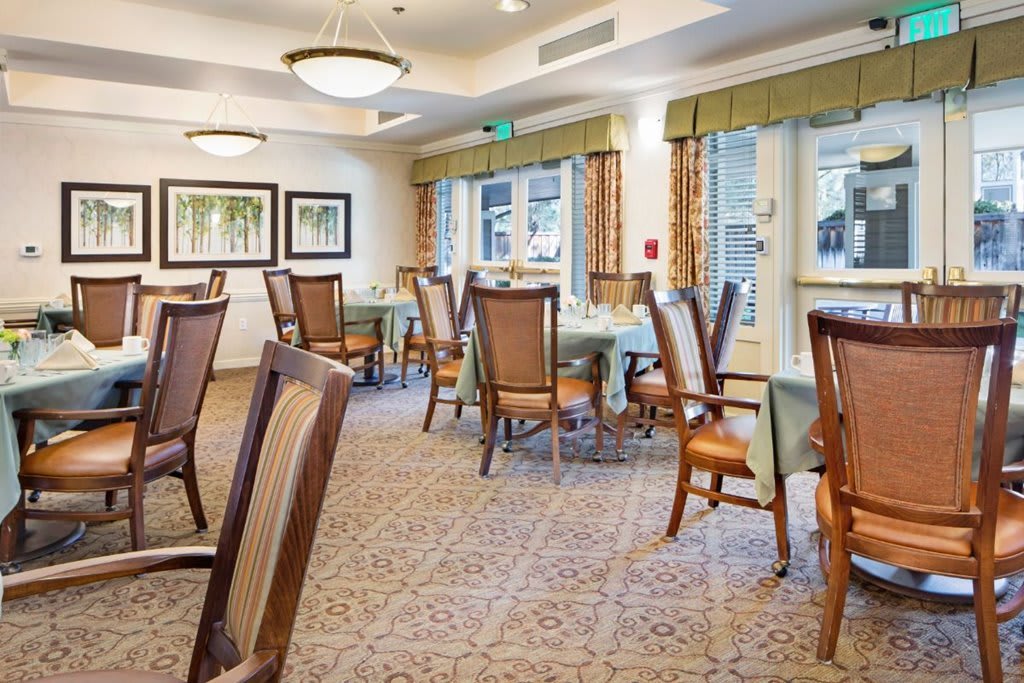 A well-lit dining room with tables and chairs arranged neatly, featuring large windows and carpeted flooring.