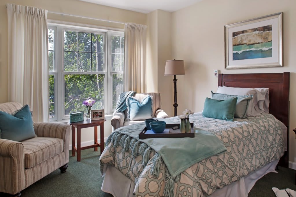 A cozy bedroom with soft lighting and comfortable furnishings, featuring a bed, a chair by a large window, and tasteful decor.