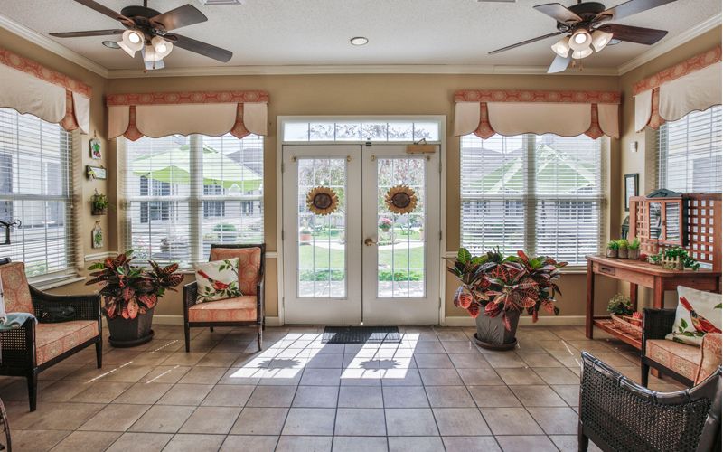 A bright living room with large windows and glass doors, featuring potted plants and comfortable seating.