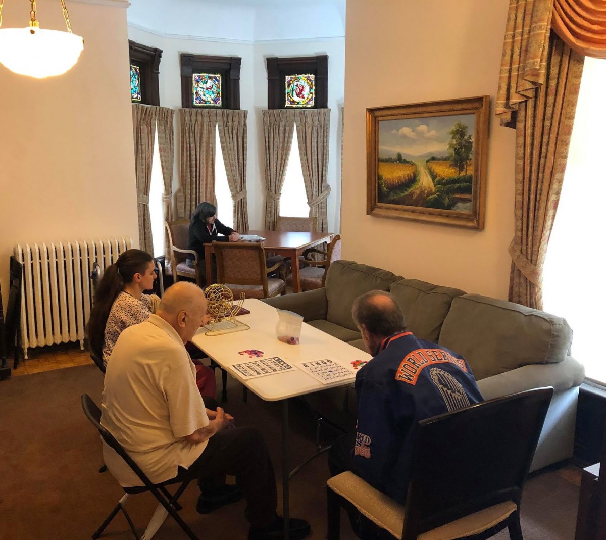 A group of people sitting at a table having a discussion in a well-lit room with curtains and a painting on the wall.