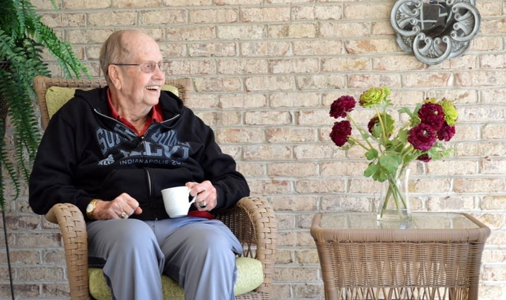 An elderly man sits comfortably with a cup of coffee, looking at flowers on a table against a brick wall.