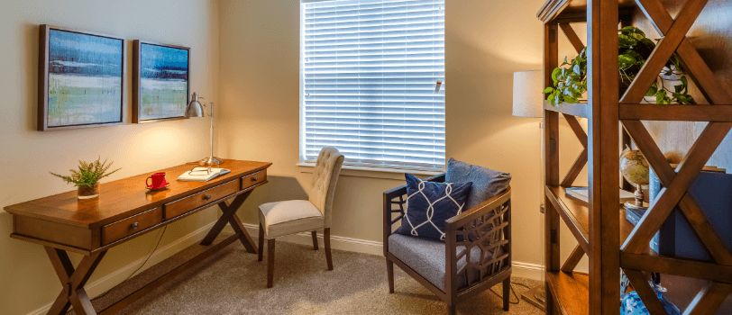 A cozy home office setup with a wooden desk, upholstered chair, and a window with blinds letting in natural light.
