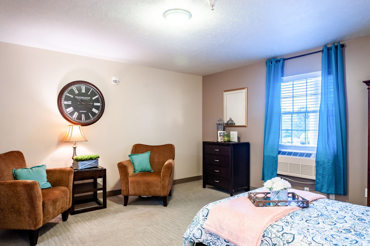 A cozy bedroom with an armchair, clock, and dresser near a window with blue curtains.