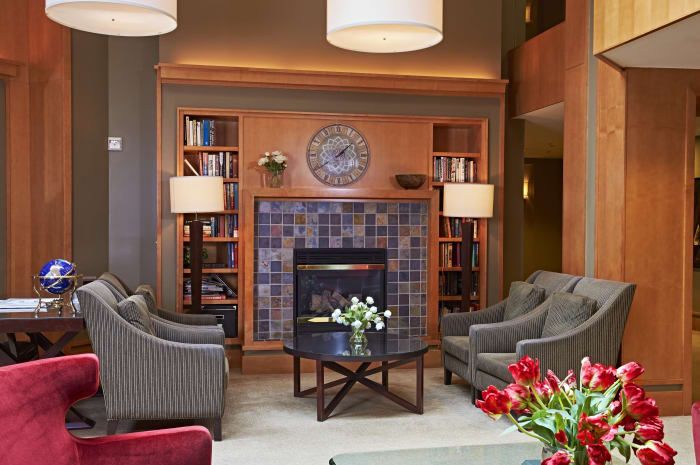A cozy living room with a fireplace, surrounded by bookshelves and armchairs, featuring a central coffee table with flowers.
