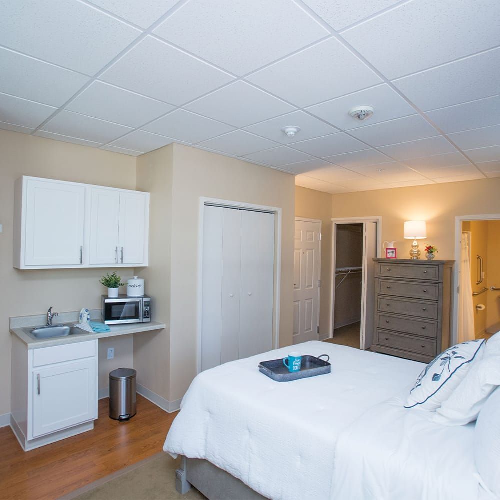 A cozy bedroom with minimalist decor, featuring a bed, dresser, and small kitchenette area.