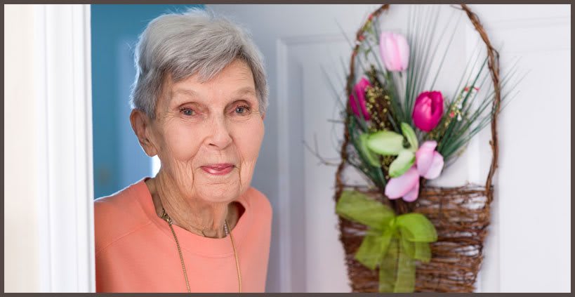 An elderly woman stands next to a floral wreath with tulips hanging on a door.