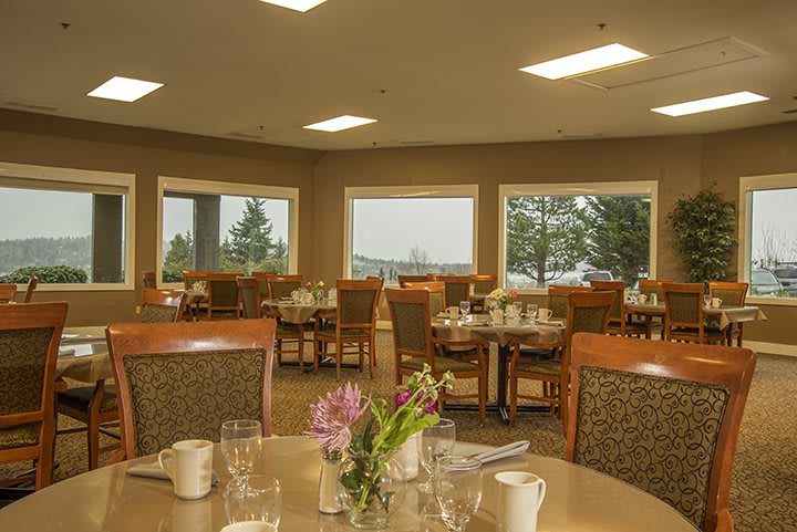 A well-lit dining room with neatly arranged tables and chairs, featuring floral centerpieces and large windows offering a scenic view.