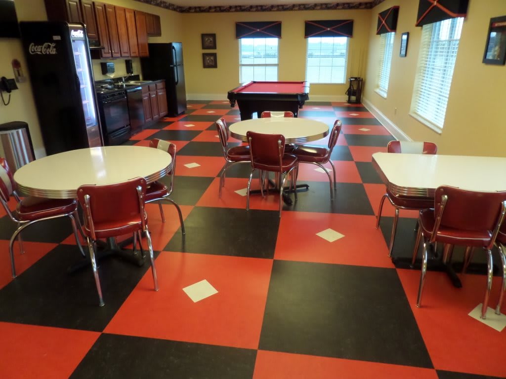 A retro-style diner interior featuring a red and black checkered floor with round tables, chairs, and a pool table.