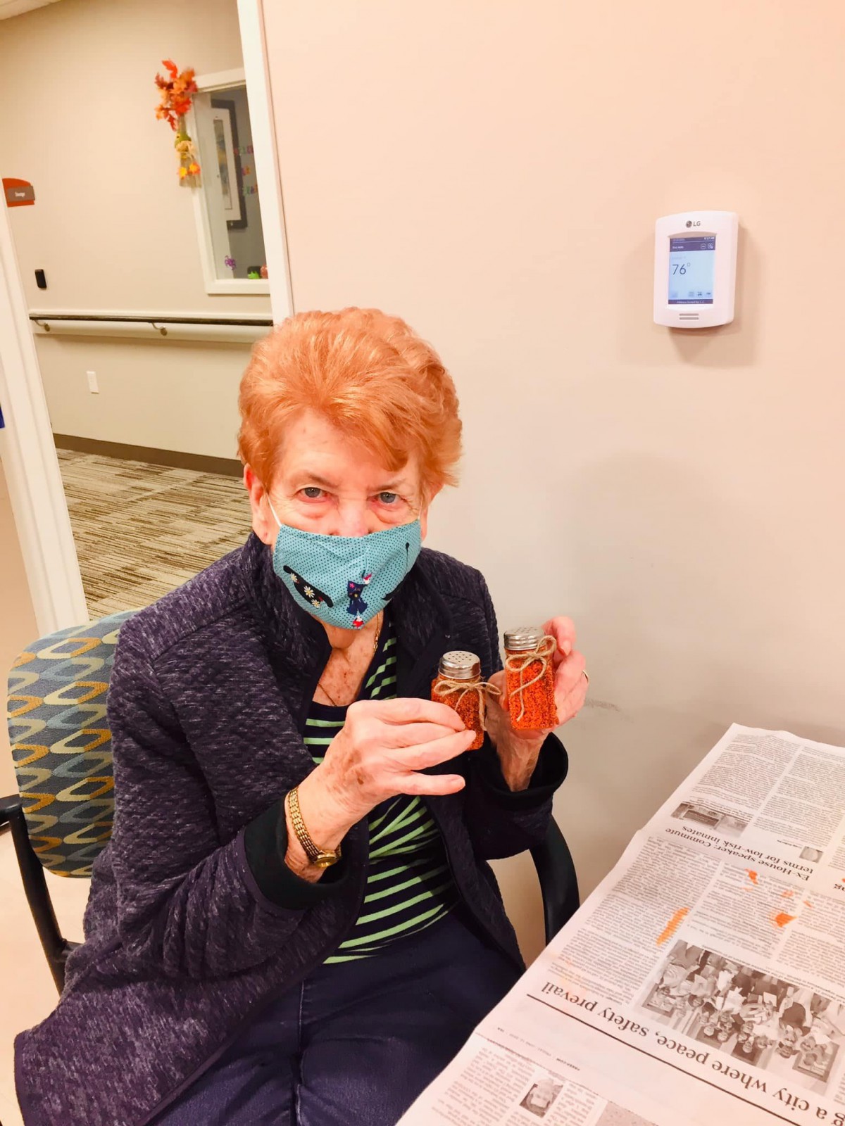A woman wearing a mask is seated indoors, holding a craft item.