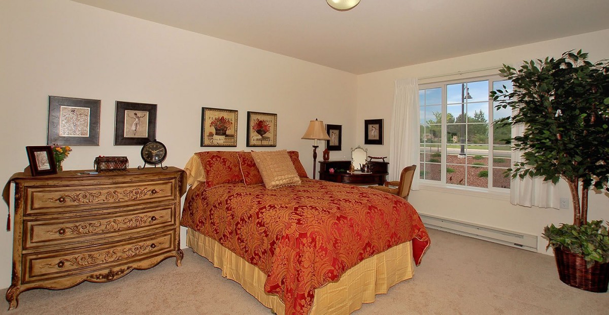 A cozy bedroom with a red and yellow bedspread, wooden furniture, and a window providing natural light.