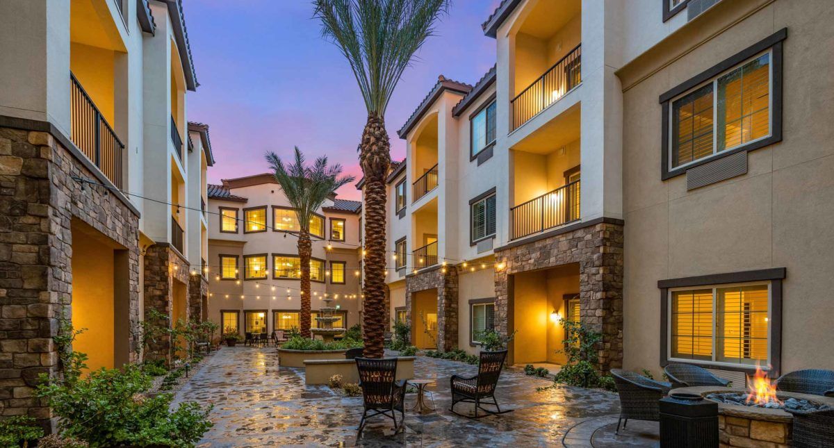 A beautifully lit apartment courtyard in the evening, featuring palm trees and outdoor seating.
