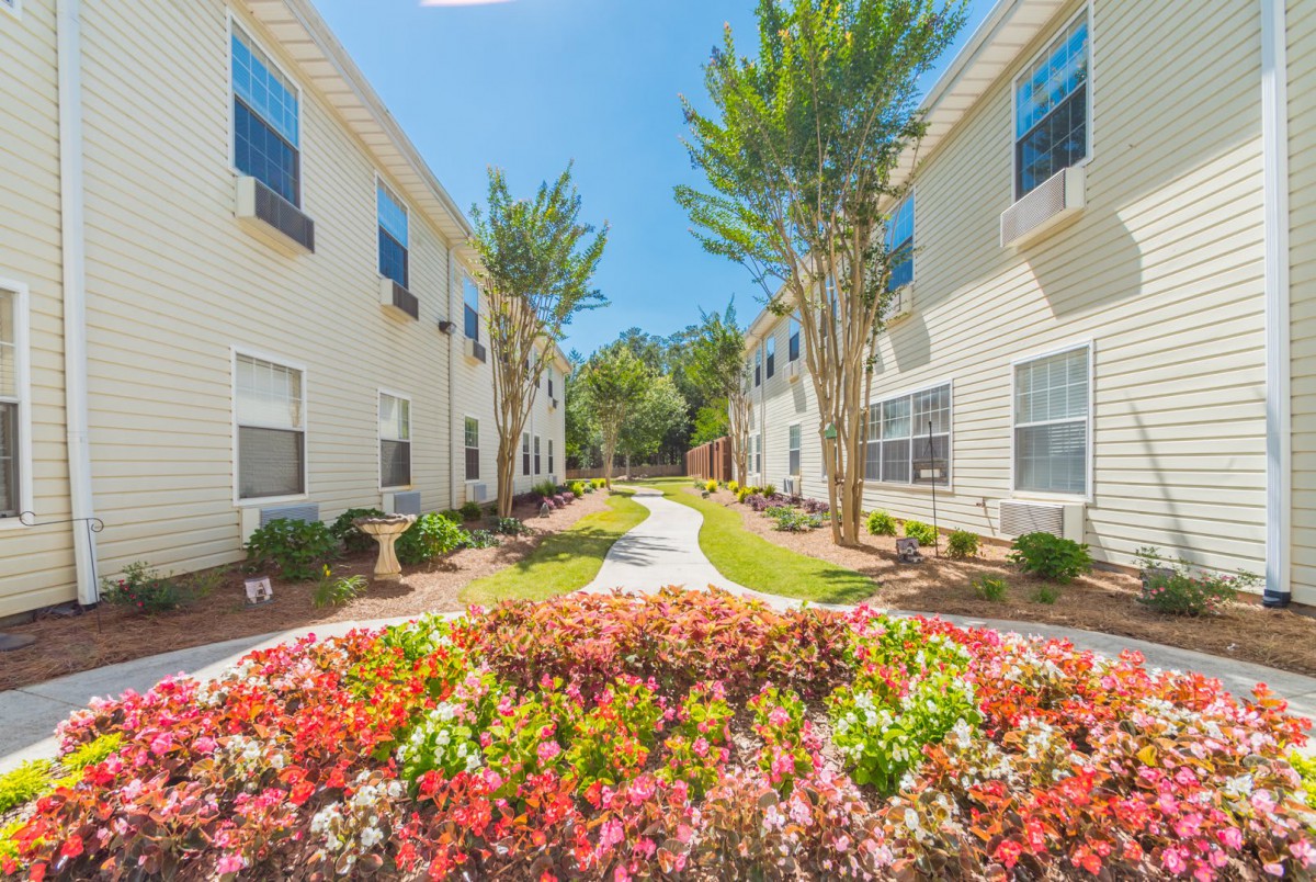 A beautifully landscaped apartment complex with colorful flowers and a central pathway under a clear blue sky.