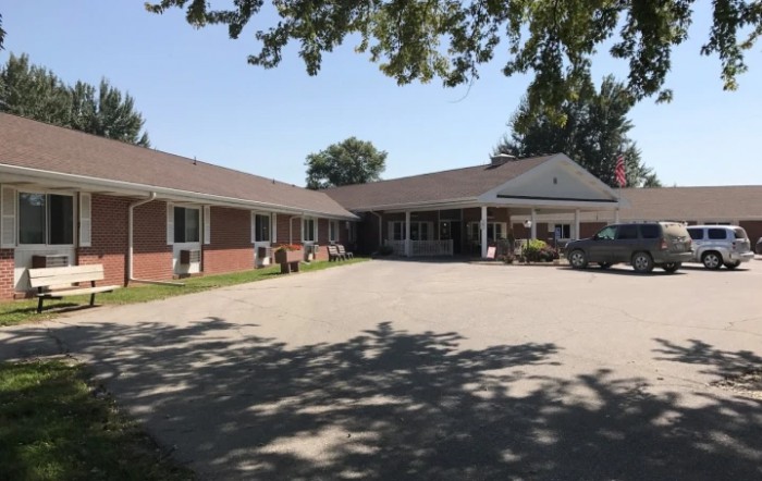 A single-story residential building with a driveway and surrounding trees casting shadows.