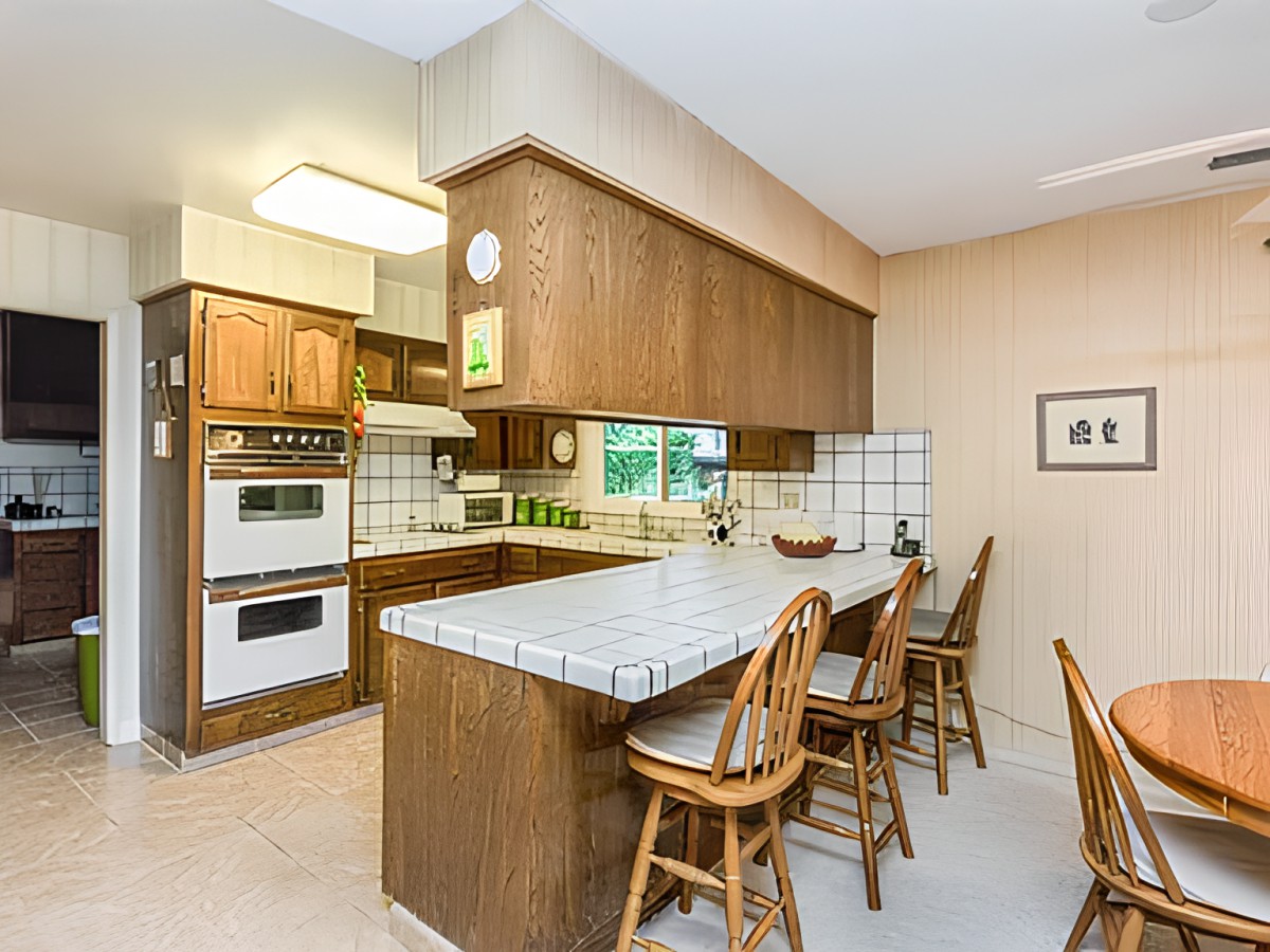 A cozy kitchen with wood cabinets and a tile countertop, featuring bar stools for counter seating.