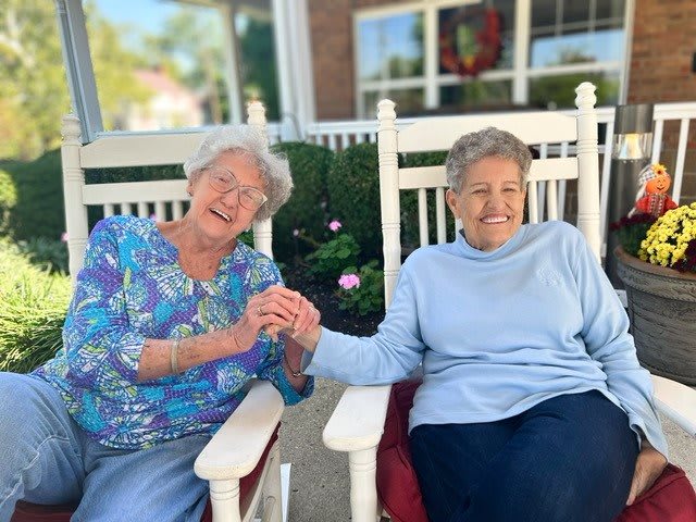 Two elderly women enjoying a moment together while sitting on rocking chairs outside.